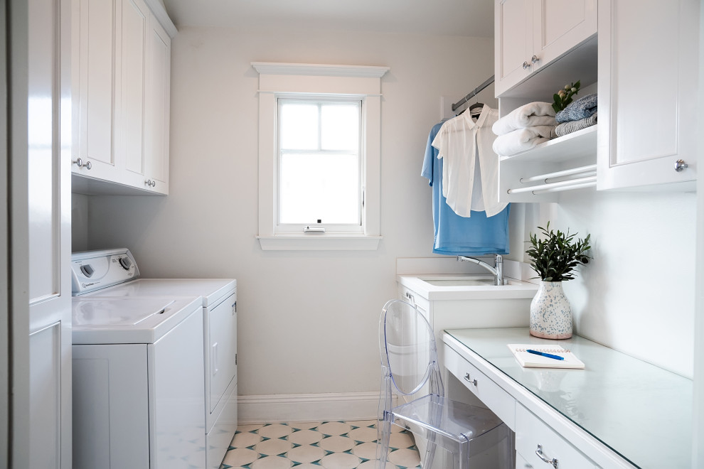 Maritim inredning av en liten vita parallell vitt tvättstuga enbart för tvätt, med en undermonterad diskho, skåp i shakerstil, vita skåp, bänkskiva i glas, vita väggar, klinkergolv i keramik, en tvättmaskin och torktumlare bredvid varandra och blått golv