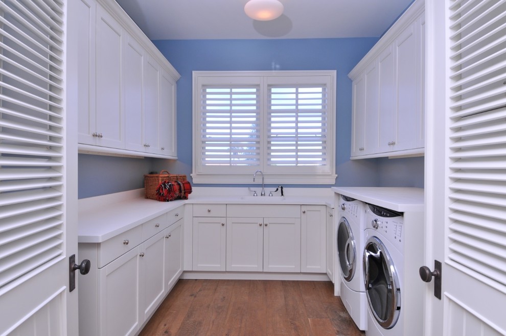 Exempel på en mellanstor maritim vita u-formad vitt tvättstuga enbart för tvätt, med en enkel diskho, blå väggar, mörkt trägolv, en tvättmaskin och torktumlare bredvid varandra, vita skåp, marmorbänkskiva, skåp i shakerstil och brunt golv