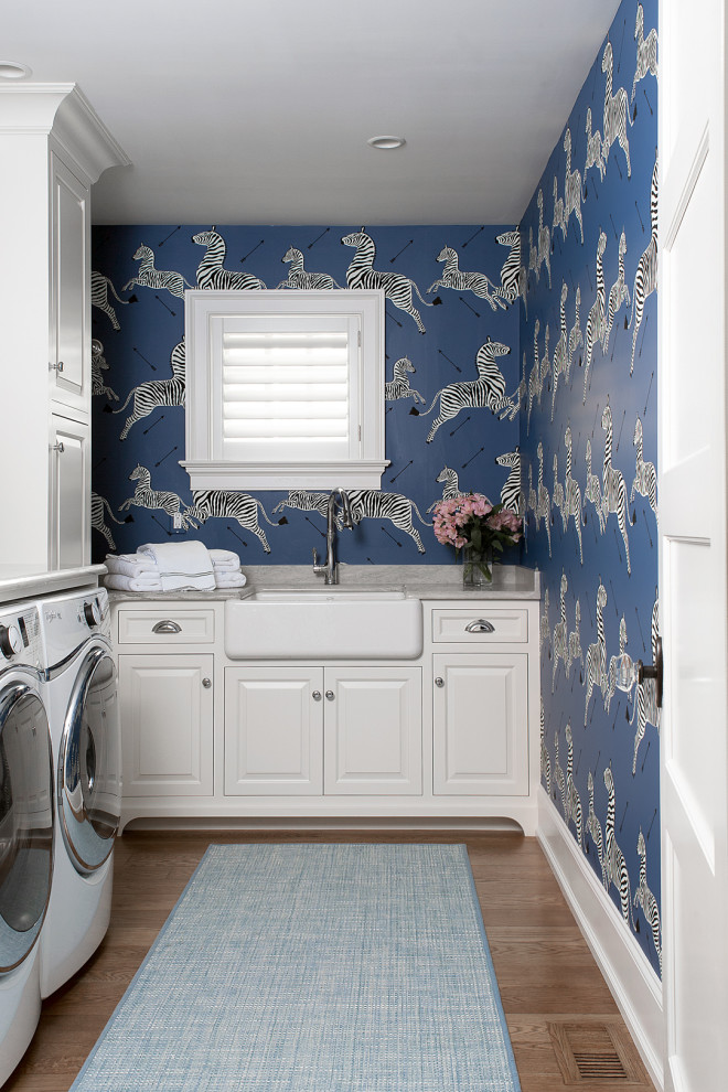 Klassisk inredning av en grå l-formad grått tvättstuga enbart för tvätt, med en rustik diskho, luckor med upphöjd panel, vita skåp, flerfärgad stänkskydd, flerfärgade väggar, mellanmörkt trägolv, en tvättmaskin och torktumlare bredvid varandra och brunt golv