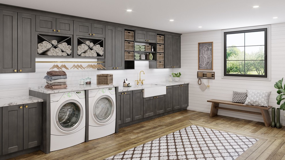Exempel på en mycket stor lantlig grå linjär grått tvättstuga enbart för tvätt, med en rustik diskho, skåp i shakerstil, grå skåp, granitbänkskiva, vita väggar, ljust trägolv, en tvättmaskin och torktumlare bredvid varandra och brunt golv