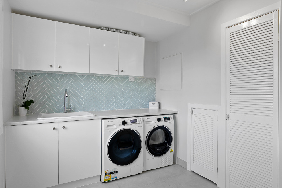 Einzeiliger Maritimer Hauswirtschaftsraum mit Einbauwaschbecken, flächenbündigen Schrankfronten, weißen Schränken, Küchenrückwand in Blau, weißer Wandfarbe, Waschmaschine und Trockner nebeneinander, grauem Boden und grauer Arbeitsplatte in Brisbane
