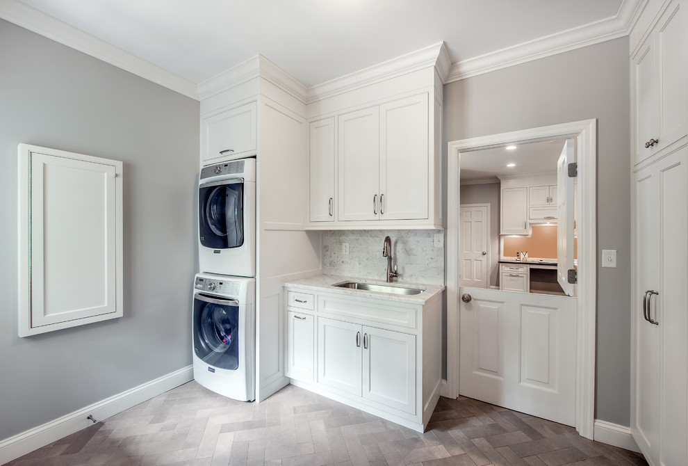 This is an example of a medium sized classic l-shaped utility room in New York with a submerged sink, beaded cabinets, white cabinets, marble worktops, grey walls, porcelain flooring and a stacked washer and dryer.