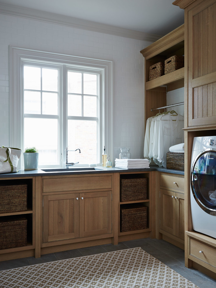 Inredning av en klassisk mellanstor l-formad tvättstuga enbart för tvätt, med en undermonterad diskho, skåp i shakerstil, skåp i mellenmörkt trä, vita väggar, en tvättmaskin och torktumlare bredvid varandra och grått golv