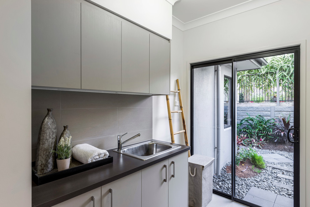 Mid-sized trendy single-wall ceramic tile dedicated laundry room photo in Brisbane with a drop-in sink, flat-panel cabinets, gray cabinets, laminate countertops and gray walls