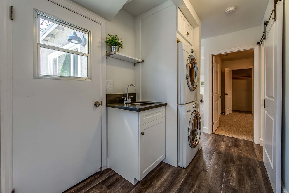 Photo of a laundry cupboard in Los Angeles with a single-bowl sink, shaker cabinets, white cabinets, white walls, medium hardwood flooring and a stacked washer and dryer.