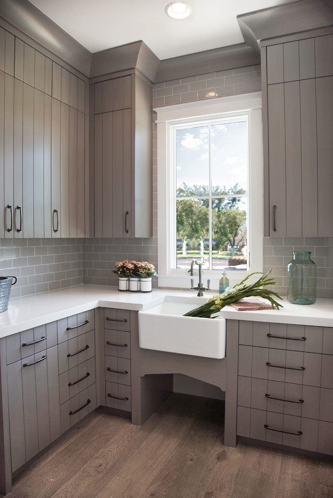 Inspiration for a medium sized traditional l-shaped separated utility room in Phoenix with a belfast sink, grey cabinets, medium hardwood flooring, flat-panel cabinets, engineered stone countertops, grey splashback, metro tiled splashback, grey walls, an integrated washer and dryer, brown floors and white worktops.