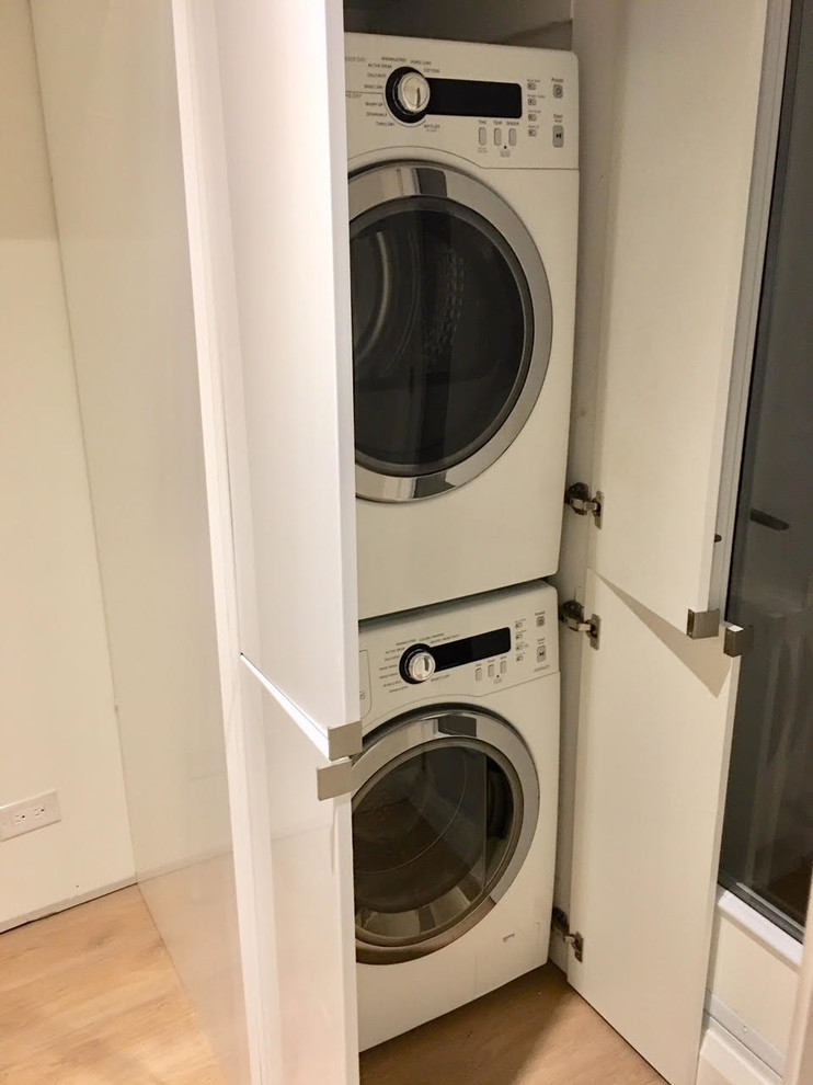 Design ideas for a small contemporary laundry cupboard in Toronto with flat-panel cabinets, white cabinets, white walls, light hardwood flooring, a concealed washer and dryer and brown floors.