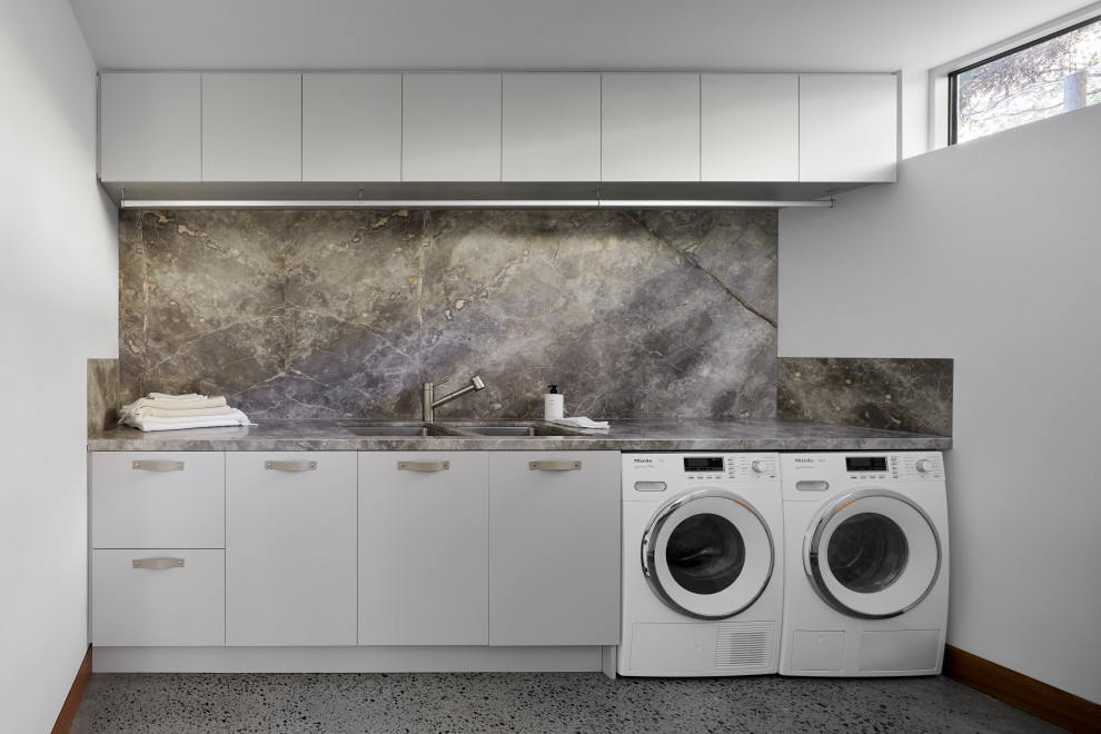 Maritim inredning av en stor grå parallell grått tvättstuga enbart för tvätt, med en dubbel diskho, grå skåp, marmorbänkskiva, grått stänkskydd, stänkskydd i marmor, vita väggar, betonggolv, en tvättmaskin och torktumlare bredvid varandra och grått golv