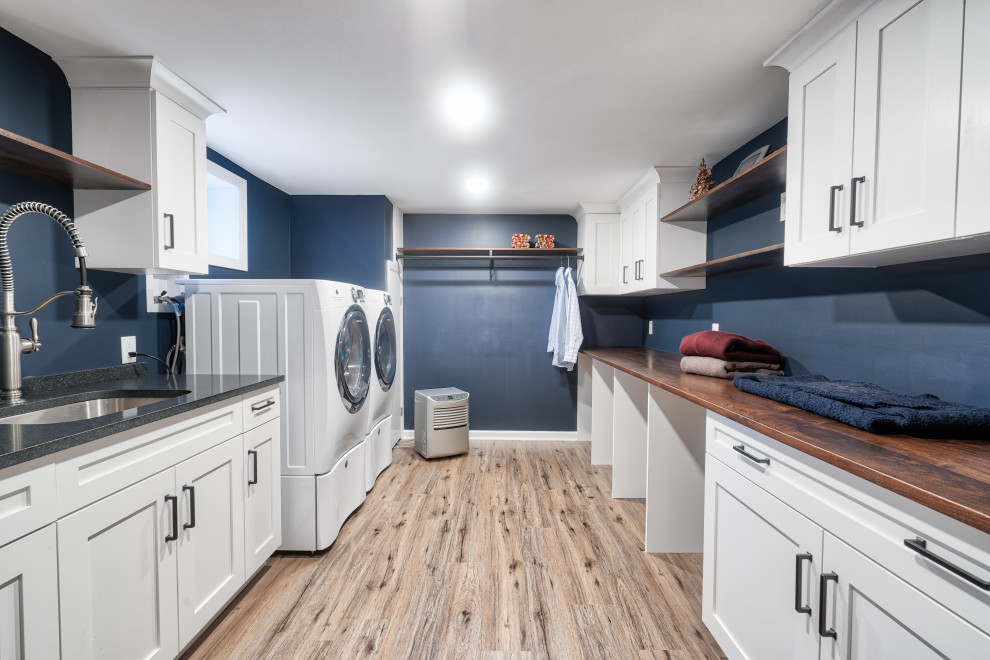 Inredning av en lantlig parallell tvättstuga enbart för tvätt, med en undermonterad diskho, skåp i shakerstil, vita skåp, blå väggar, ljust trägolv och en tvättmaskin och torktumlare bredvid varandra