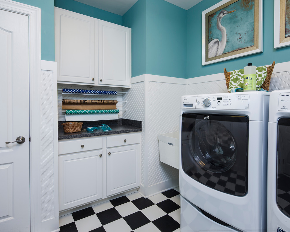 Modelo de lavadero multiusos costero con pila para lavar, armarios con paneles con relieve, puertas de armario blancas, paredes azules y lavadora y secadora juntas