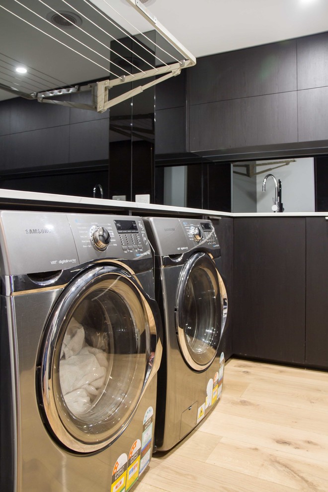 Medium sized modern l-shaped separated utility room in Melbourne with a built-in sink, flat-panel cabinets, black cabinets, black walls, medium hardwood flooring, a side by side washer and dryer, brown floors and white worktops.