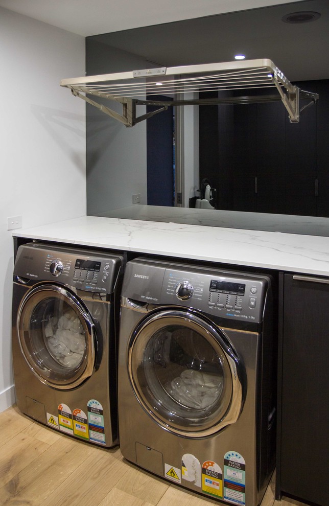 Medium sized modern l-shaped separated utility room in Melbourne with a built-in sink, flat-panel cabinets, black cabinets, medium hardwood flooring, a side by side washer and dryer, brown floors, white worktops and black walls.
