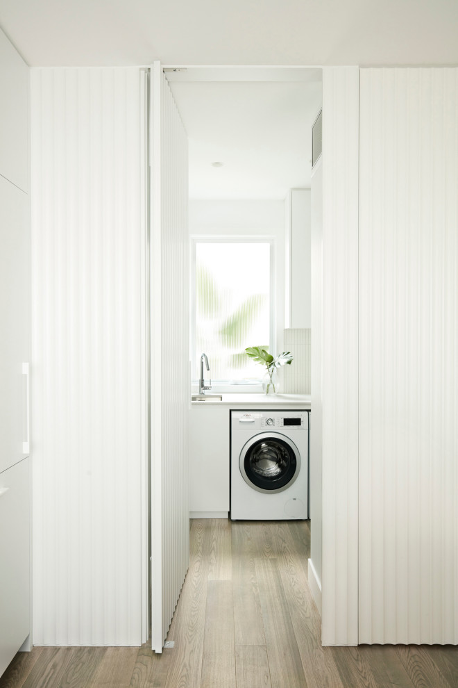 Small trendy l-shaped medium tone wood floor and brown floor dedicated laundry room photo in Melbourne with a single-bowl sink, flat-panel cabinets, white cabinets, quartz countertops, white backsplash, ceramic backsplash, white walls, a side-by-side washer/dryer and gray countertops