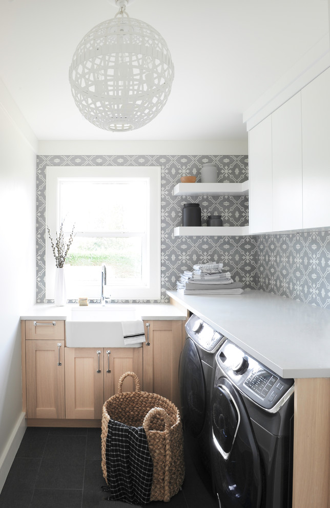 Bild på en lantlig grå l-formad grått tvättstuga enbart för tvätt, med en rustik diskho, skåp i shakerstil, skåp i ljust trä, flerfärgade väggar, en tvättmaskin och torktumlare bredvid varandra och svart golv