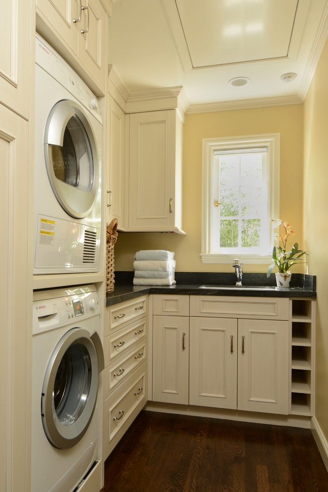 Ejemplo de lavadero tradicional con lavadora y secadora apiladas, puertas de armario blancas, paredes amarillas y encimeras negras