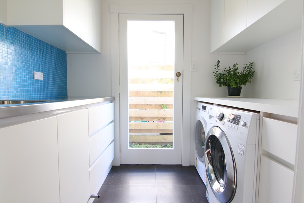 オークランドにあるお手頃価格の中くらいなモダンスタイルのおしゃれな洗濯室 (ll型、フラットパネル扉のキャビネット、白いキャビネット、ステンレスカウンター、青いキッチンパネル、ガラスタイルのキッチンパネル、左右配置の洗濯機・乾燥機、白いキッチンカウンター) の写真