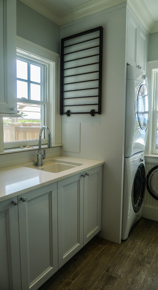 Photo of a medium sized classic single-wall separated utility room in Boston with a built-in sink, grey walls, medium hardwood flooring, a stacked washer and dryer, recessed-panel cabinets, white cabinets, quartz worktops and white worktops.