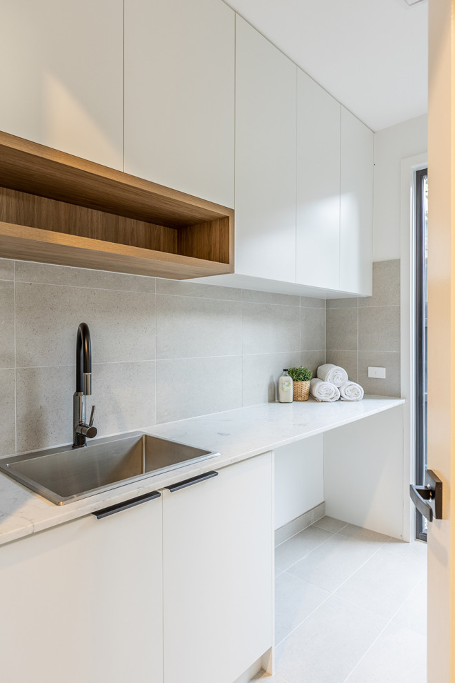 Inspiration för moderna linjära vitt tvättstugor enbart för tvätt, med en enkel diskho, vita skåp, marmorbänkskiva, grått stänkskydd, stänkskydd i porslinskakel, grå väggar, klinkergolv i porslin, en tvättmaskin och torktumlare bredvid varandra och grått golv