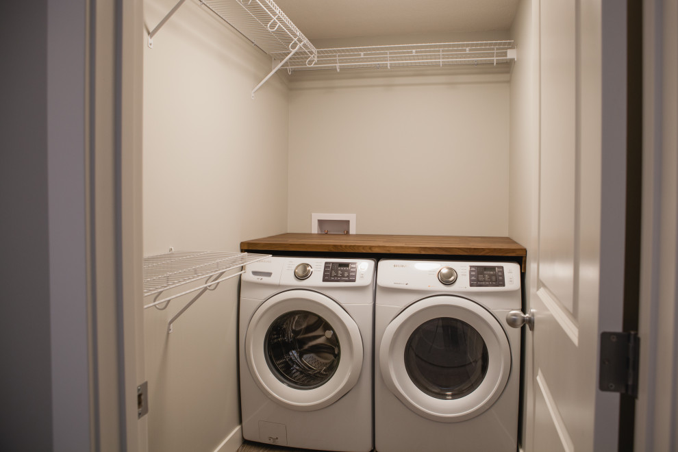Einzeiliger, Kleiner Moderner Hauswirtschaftsraum mit Waschmaschinenschrank, Arbeitsplatte aus Holz, beiger Wandfarbe, Waschmaschine und Trockner nebeneinander und brauner Arbeitsplatte in Calgary