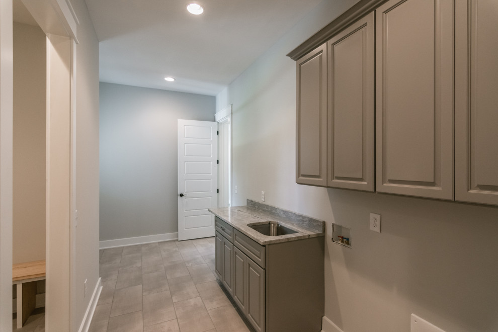 Photo of a medium sized country single-wall utility room in Nashville with a submerged sink, raised-panel cabinets, grey cabinets, granite worktops, grey walls, porcelain flooring, a side by side washer and dryer, beige floors and grey worktops.