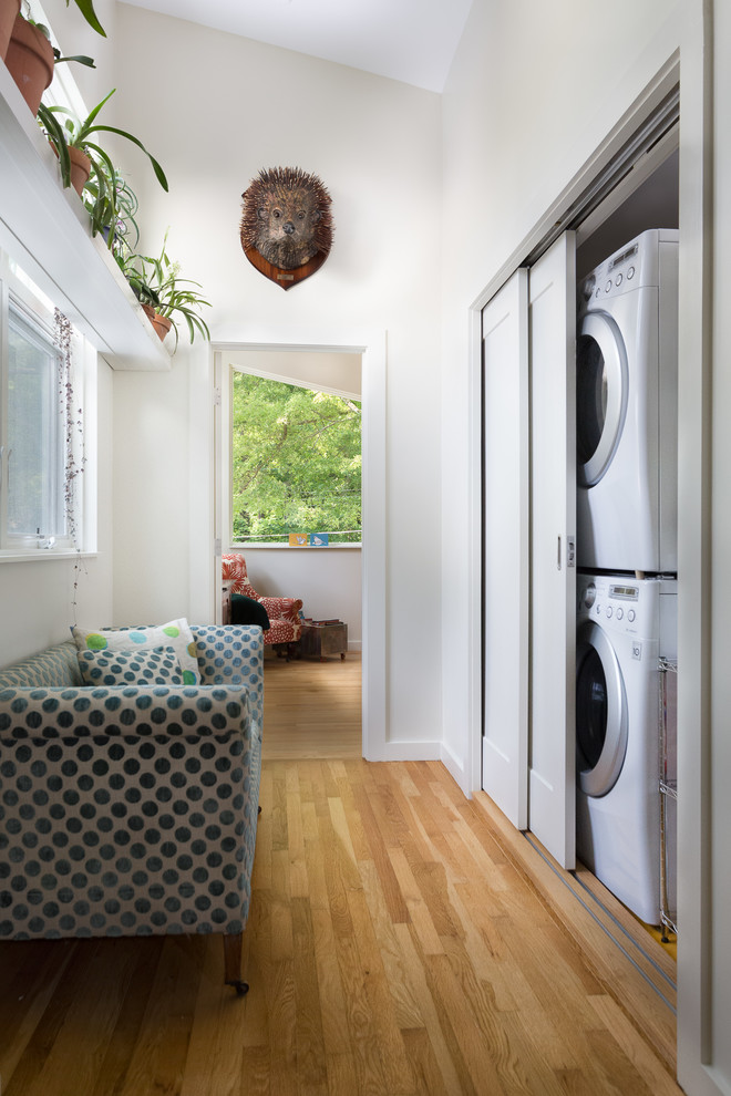 Diseño de armario lavadero lineal actual pequeño con paredes blancas, suelo de madera clara y lavadora y secadora apiladas