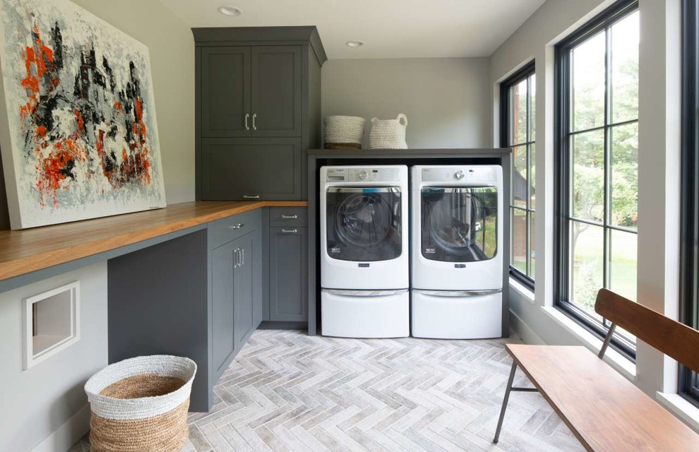 Inredning av en skandinavisk bruna l-formad brunt tvättstuga enbart för tvätt, med grå skåp, träbänkskiva, klinkergolv i porslin, en tvättmaskin och torktumlare bredvid varandra, grått golv, skåp i shakerstil och grå väggar