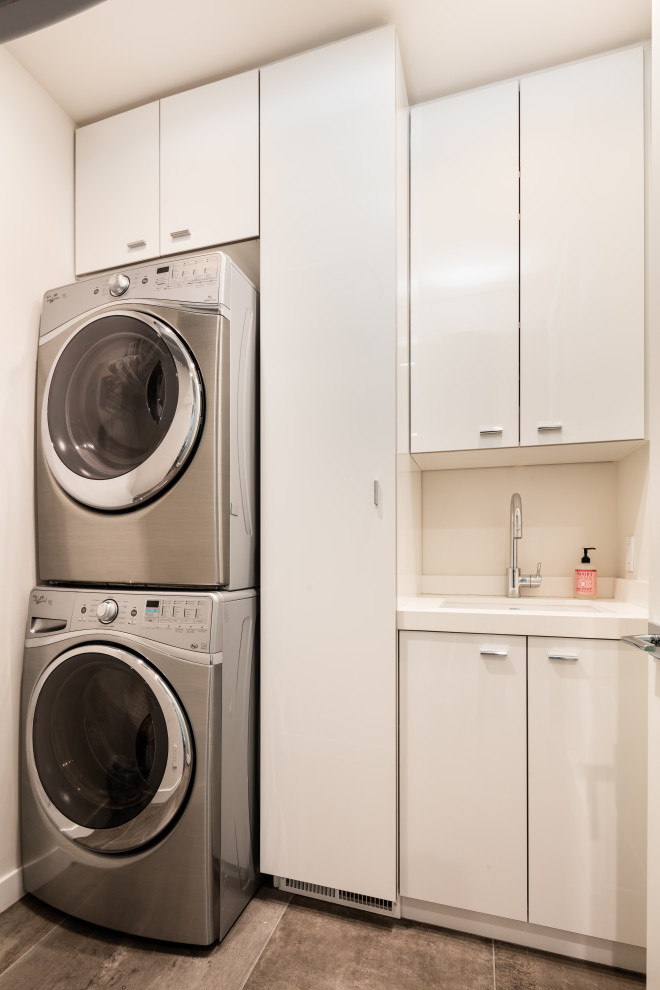 This is an example of a small contemporary single-wall separated utility room in Orange County with a submerged sink, flat-panel cabinets, white cabinets, engineered stone countertops, white walls, porcelain flooring, a stacked washer and dryer, grey floors and white worktops.