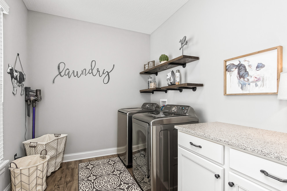 Inredning av en lantlig grå linjär grått tvättstuga enbart för tvätt, med luckor med infälld panel, vita skåp, grå väggar, mörkt trägolv, en tvättmaskin och torktumlare bredvid varandra och brunt golv