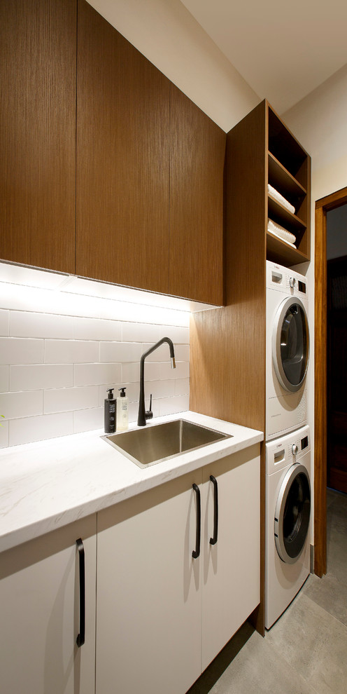 Imagen de cuarto de lavado lineal contemporáneo de tamaño medio con fregadero encastrado, armarios con paneles lisos, encimera de cuarzo compacto, paredes blancas, lavadora y secadora apiladas y puertas de armario de madera oscura