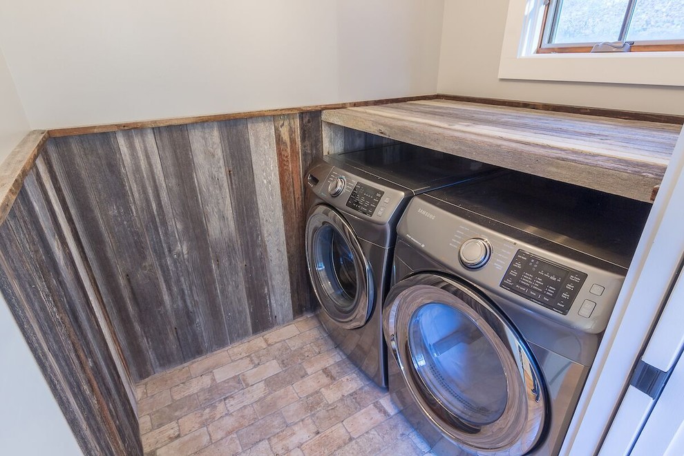 Modelo de cuarto de lavado rural pequeño con puertas de armario con efecto envejecido, paredes grises, suelo de ladrillo y lavadora y secadora juntas