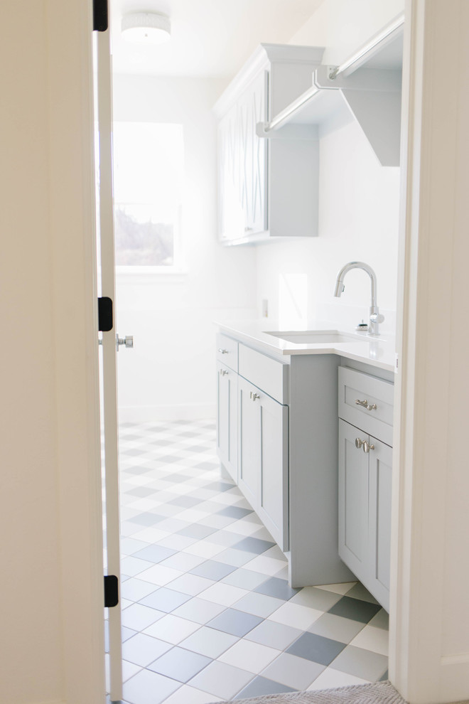 Bild på en mellanstor lantlig vita linjär vitt tvättstuga enbart för tvätt, med en undermonterad diskho, skåp i shakerstil, blå skåp, bänkskiva i kvarts, vita väggar, klinkergolv i porslin, en tvättmaskin och torktumlare bredvid varandra och grått golv