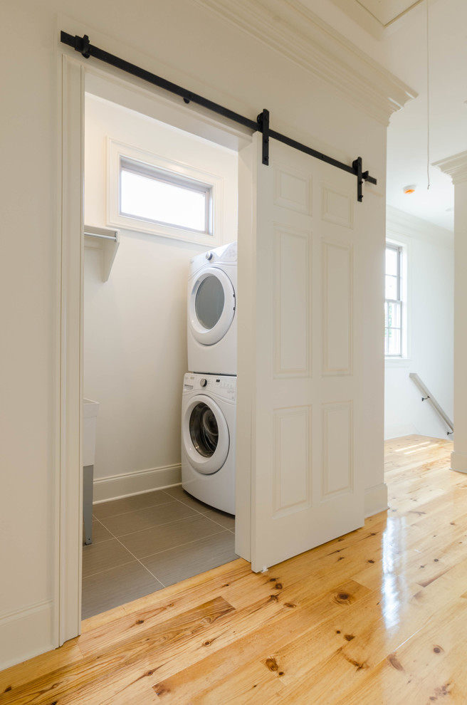 Modelo de cuarto de lavado tradicional con pila para lavar, paredes blancas, suelo de madera clara, lavadora y secadora apiladas y suelo marrón
