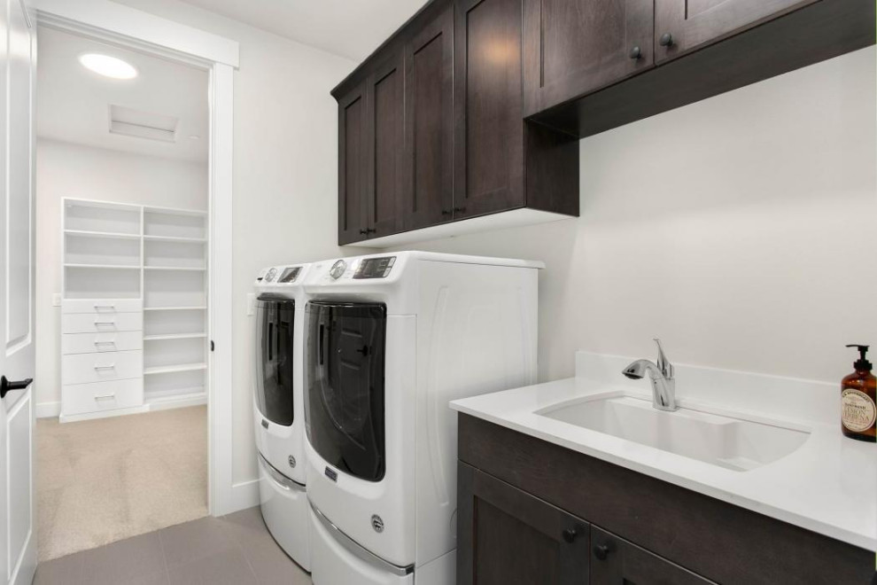 Modern inredning av en stor vita l-formad vitt tvättstuga enbart för tvätt, med en nedsänkt diskho, skåp i shakerstil, skåp i mörkt trä, bänkskiva i kvarts, vita väggar, klinkergolv i porslin, en tvättmaskin och torktumlare bredvid varandra och grått golv