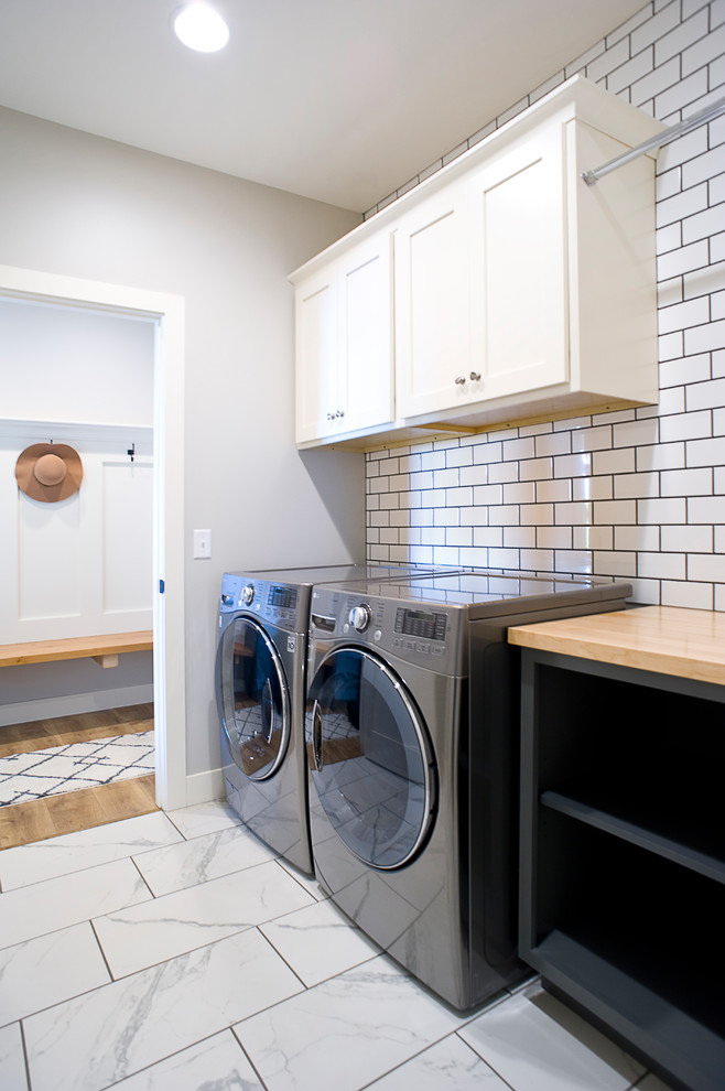 Example of a mid-sized transitional single-wall ceramic tile and white floor dedicated laundry room design in Other with shaker cabinets, white cabinets, wood countertops, gray walls, a side-by-side washer/dryer and brown countertops