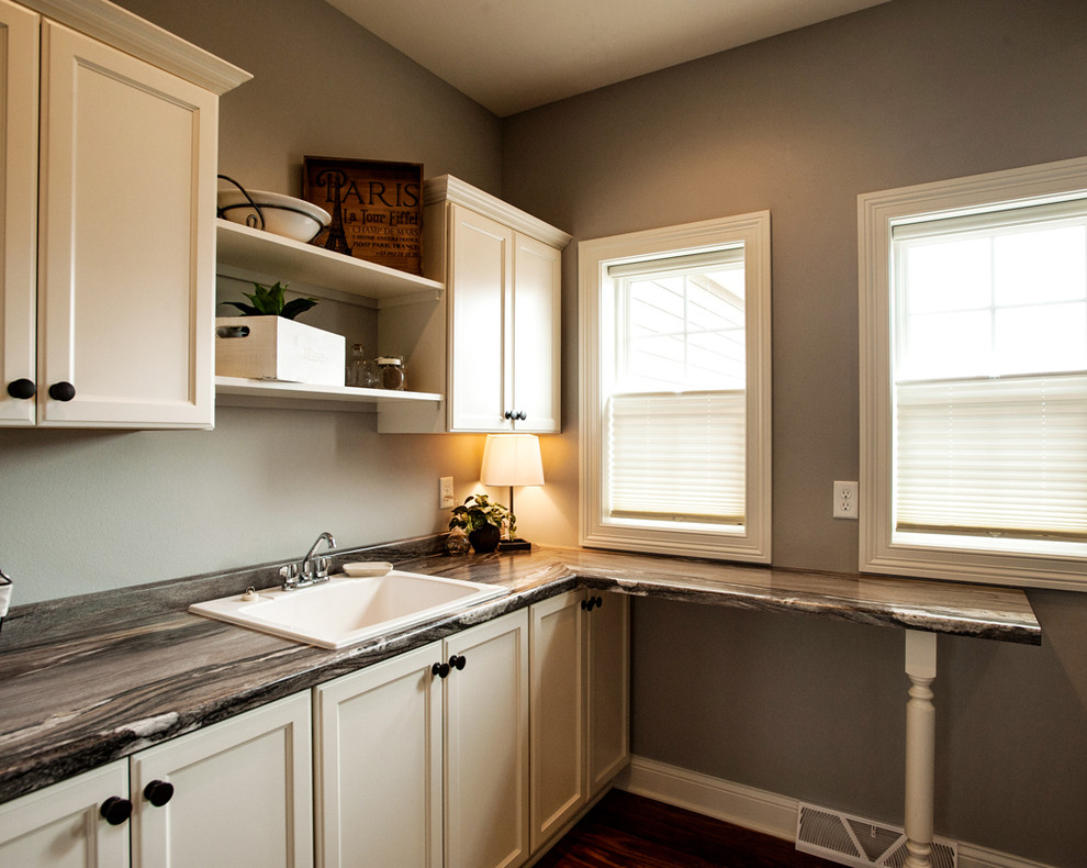 Klassisk inredning av en stor grå l-formad grått tvättstuga enbart för tvätt, med luckor med infälld panel, vita skåp, granitbänkskiva, en nedsänkt diskho, grå väggar och mörkt trägolv
