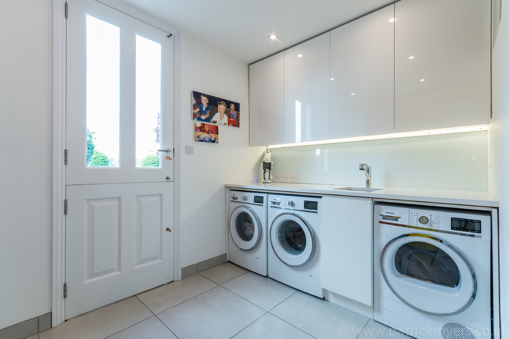 Inspiration for a medium sized contemporary separated utility room in London with a single-bowl sink, flat-panel cabinets, white cabinets, white walls, a side by side washer and dryer and white floors.
