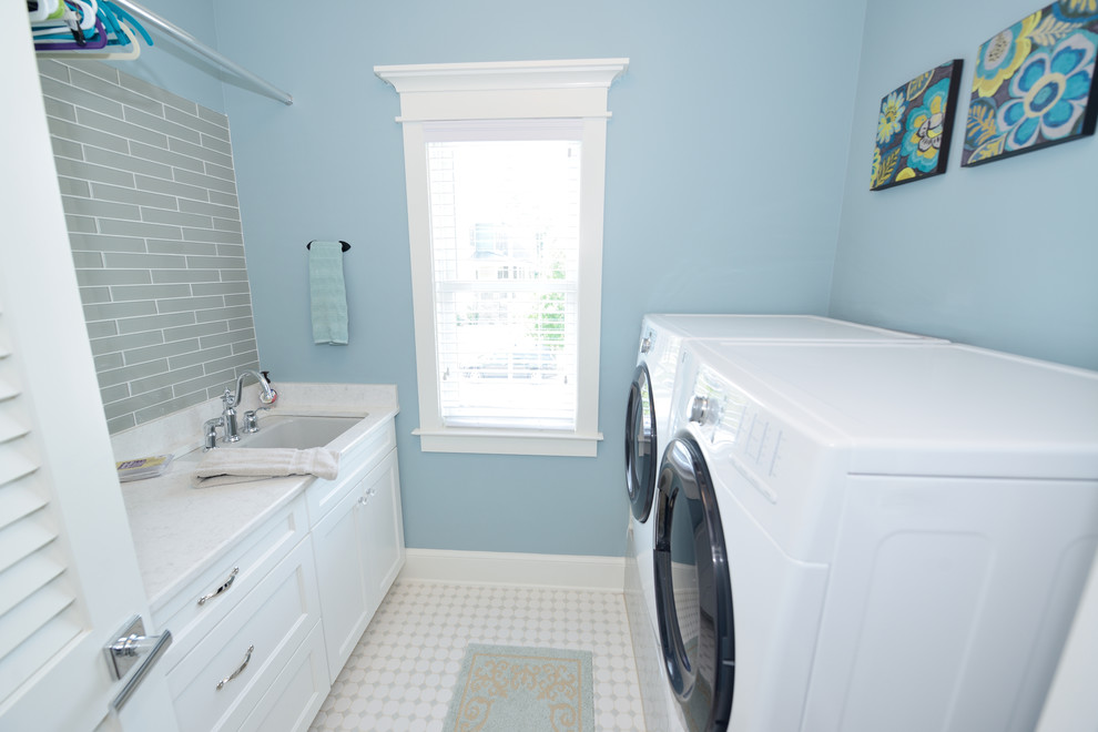 Klassisk inredning av en liten parallell tvättstuga enbart för tvätt, med vita skåp, blå väggar, en tvättmaskin och torktumlare bredvid varandra, en undermonterad diskho, skåp i shakerstil, bänkskiva i täljsten, klinkergolv i keramik och vitt golv