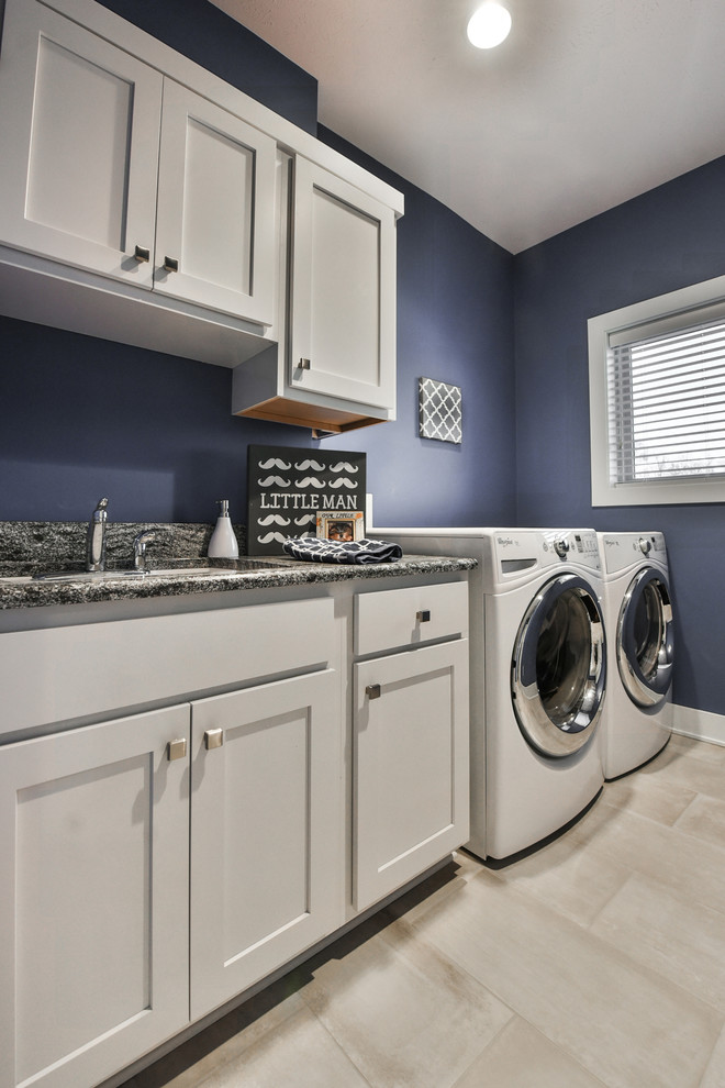 Exempel på en mellanstor klassisk grå linjär grått tvättstuga enbart för tvätt, med en undermonterad diskho, skåp i shakerstil, vita skåp, granitbänkskiva, lila väggar, klinkergolv i porslin, en tvättmaskin och torktumlare bredvid varandra och beiget golv