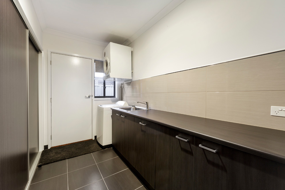 This is an example of a medium sized modern galley utility room in Sydney with a single-bowl sink, flat-panel cabinets, grey cabinets, laminate countertops, white walls, ceramic flooring and a stacked washer and dryer.