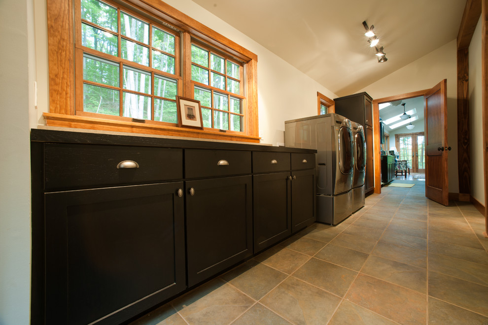 Foto de cuarto de lavado lineal clásico grande con armarios estilo shaker, puertas de armario negras, paredes blancas, suelo de pizarra y lavadora y secadora juntas