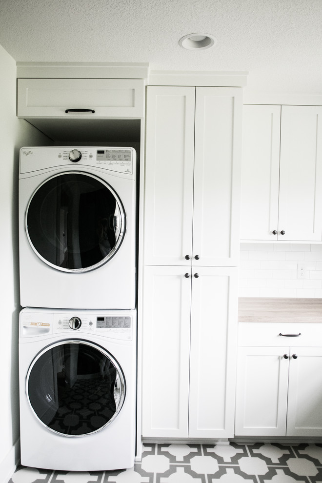 Imagen de cuarto de lavado en L de estilo de casa de campo grande con fregadero encastrado, armarios con paneles lisos, puertas de armario blancas, encimera de laminado, suelo vinílico, lavadora y secadora apiladas y paredes grises