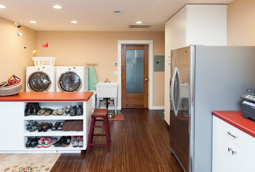 ポートランドにあるお手頃価格の中くらいなエクレクティックスタイルのおしゃれな家事室 (ll型、スロップシンク、フラットパネル扉のキャビネット、白いキャビネット、ラミネートカウンター、オレンジの壁、竹フローリング、左右配置の洗濯機・乾燥機) の写真