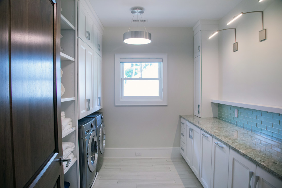 ミルウォーキーにある高級な中くらいなビーチスタイルのおしゃれな洗濯室 (ll型、落し込みパネル扉のキャビネット、白いキャビネット、御影石カウンター、青いキッチンパネル、磁器タイルのキッチンパネル、ベージュの壁、磁器タイルの床、左右配置の洗濯機・乾燥機、ベージュの床、ベージュのキッチンカウンター) の写真