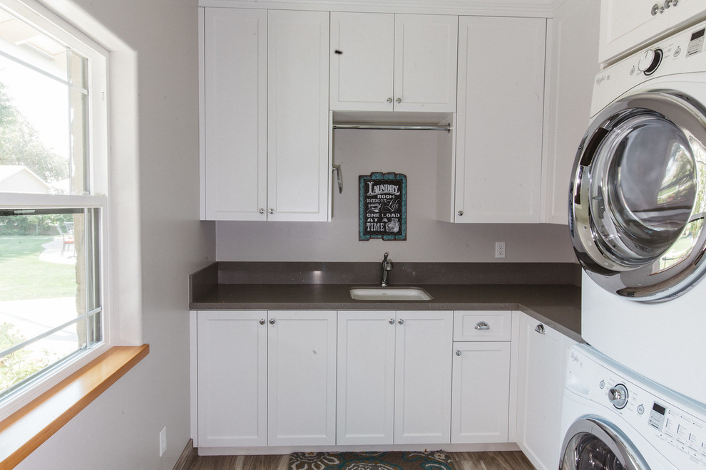 Large farmhouse l-shaped separated utility room in Boston with a submerged sink, white cabinets, composite countertops, grey walls, a stacked washer and dryer, shaker cabinets and plywood flooring.