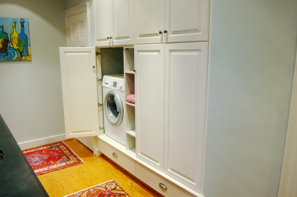 Small classic galley utility room in Richmond with raised-panel cabinets, white cabinets, blue walls, medium hardwood flooring, a concealed washer and dryer and brown floors.