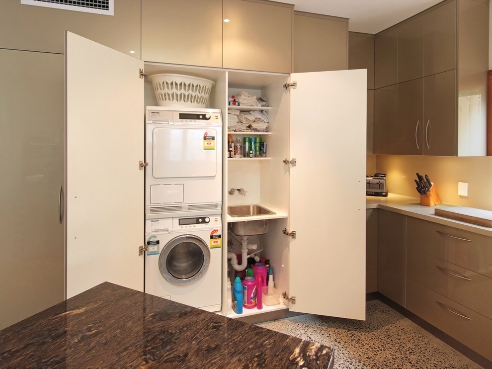 Kleiner, Einzeiliger Moderner Hauswirtschaftsraum mit Einbauwaschbecken, flächenbündigen Schrankfronten, beigen Schränken, Waschmaschine und Trockner gestapelt, Waschmaschinenschrank und Betonboden in Brisbane