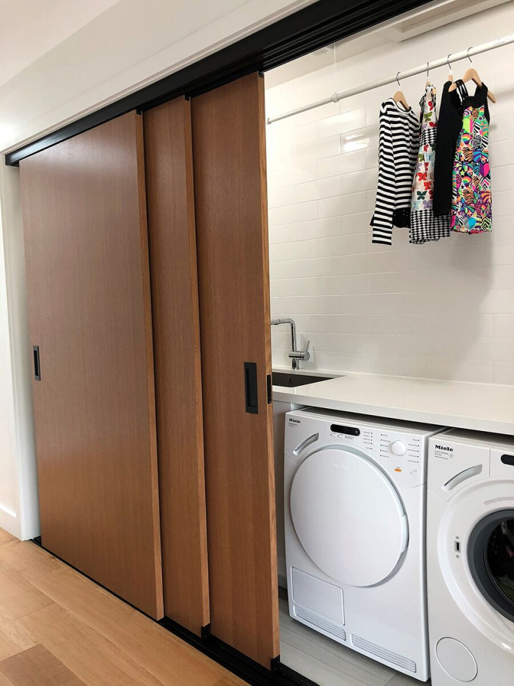 Diseño de armario lavadero moderno con fregadero integrado, armarios abiertos, puertas de armario blancas, suelo de madera en tonos medios, lavadora y secadora juntas, suelo marrón y encimeras blancas