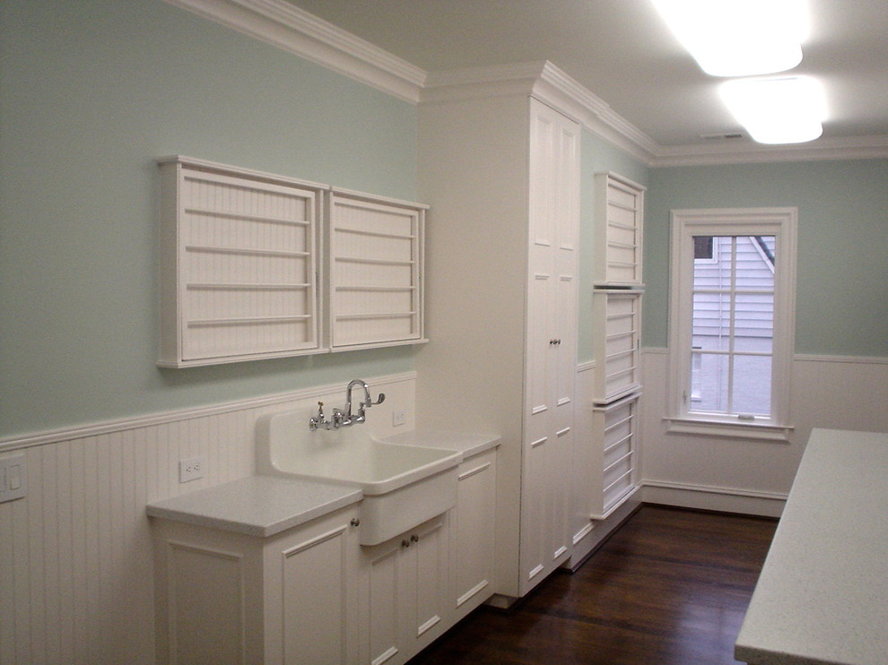 Klassisk inredning av en mellanstor vita u-formad vitt tvättstuga enbart för tvätt, med en allbänk, luckor med profilerade fronter, vita skåp, bänkskiva i kvarts, blå väggar, mörkt trägolv, en tvättmaskin och torktumlare bredvid varandra och brunt golv