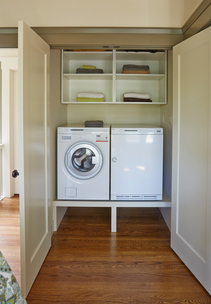 Diseño de armario lavadero lineal tradicional de tamaño medio con armarios abiertos, puertas de armario blancas, paredes blancas, suelo de madera en tonos medios y lavadora y secadora juntas