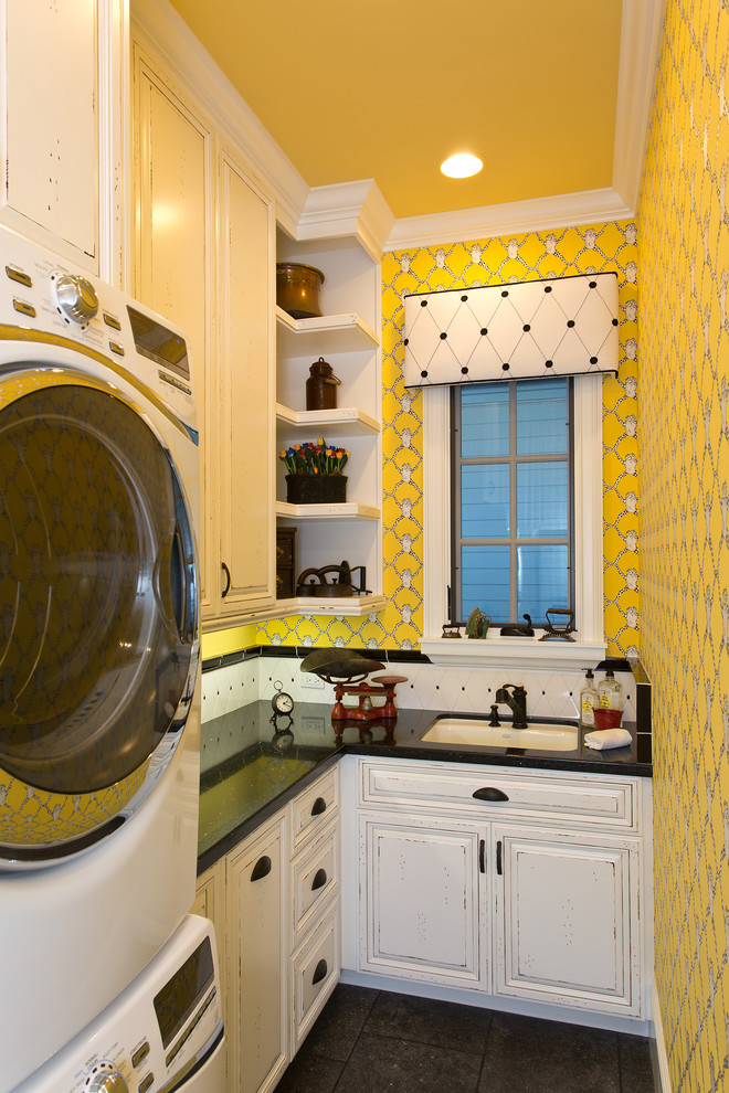 オレンジカウンティにある中くらいなトラディショナルスタイルのおしゃれな洗濯室 (L型、アンダーカウンターシンク、レイズドパネル扉のキャビネット、ヴィンテージ仕上げキャビネット、黄色い壁、上下配置の洗濯機・乾燥機、クオーツストーンカウンター、ライムストーンの床、黒い床、黒いキッチンカウンター) の写真